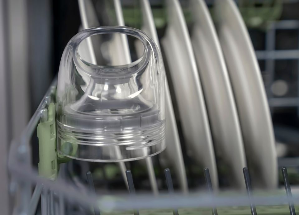 A close-up of a dishwasher rack with a transparent baby bottle nipple attachment beside several white plates.