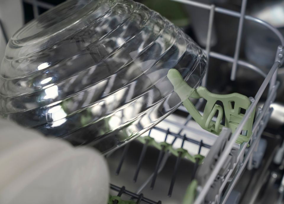 A glass bowl is securely held in place by a green clip on the dishwasher rack, preventing movement during wash.