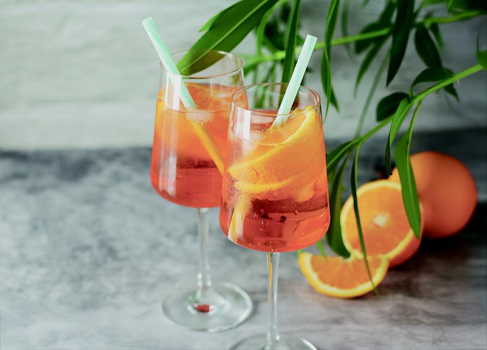Bicchiere di Spritz con ghiaccio e fetta d'arancia. Mezze arance accanto. Sfondo chiaro minimalista.
